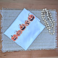 A folded white tshirt with a printed row of orange jack-o'-lantern pumpkins sits on a woven placemat on a wooden table, next to a string of light wood beads.