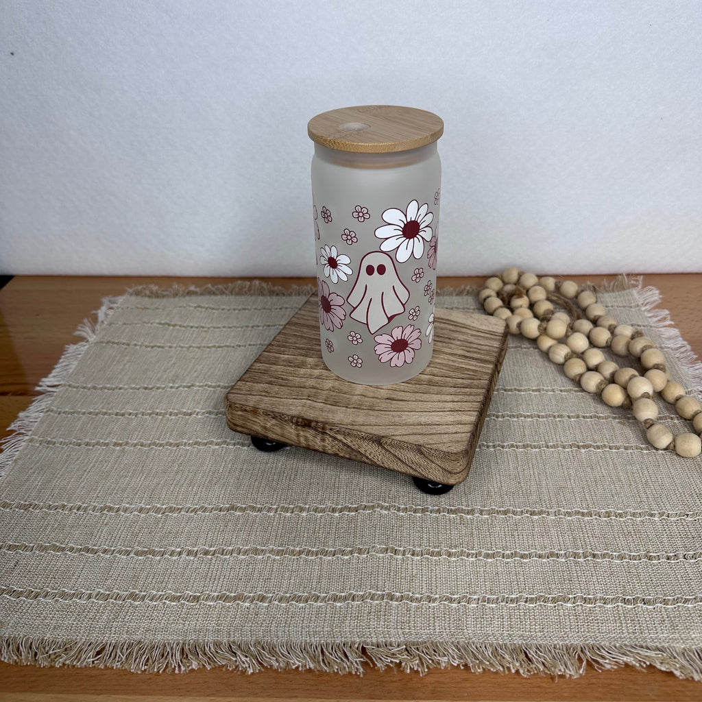 A frosted glass cup with a light wood lid, decorated with a cute ghost and daisy pattern, sitting on a wooden riser on a woven placemat.