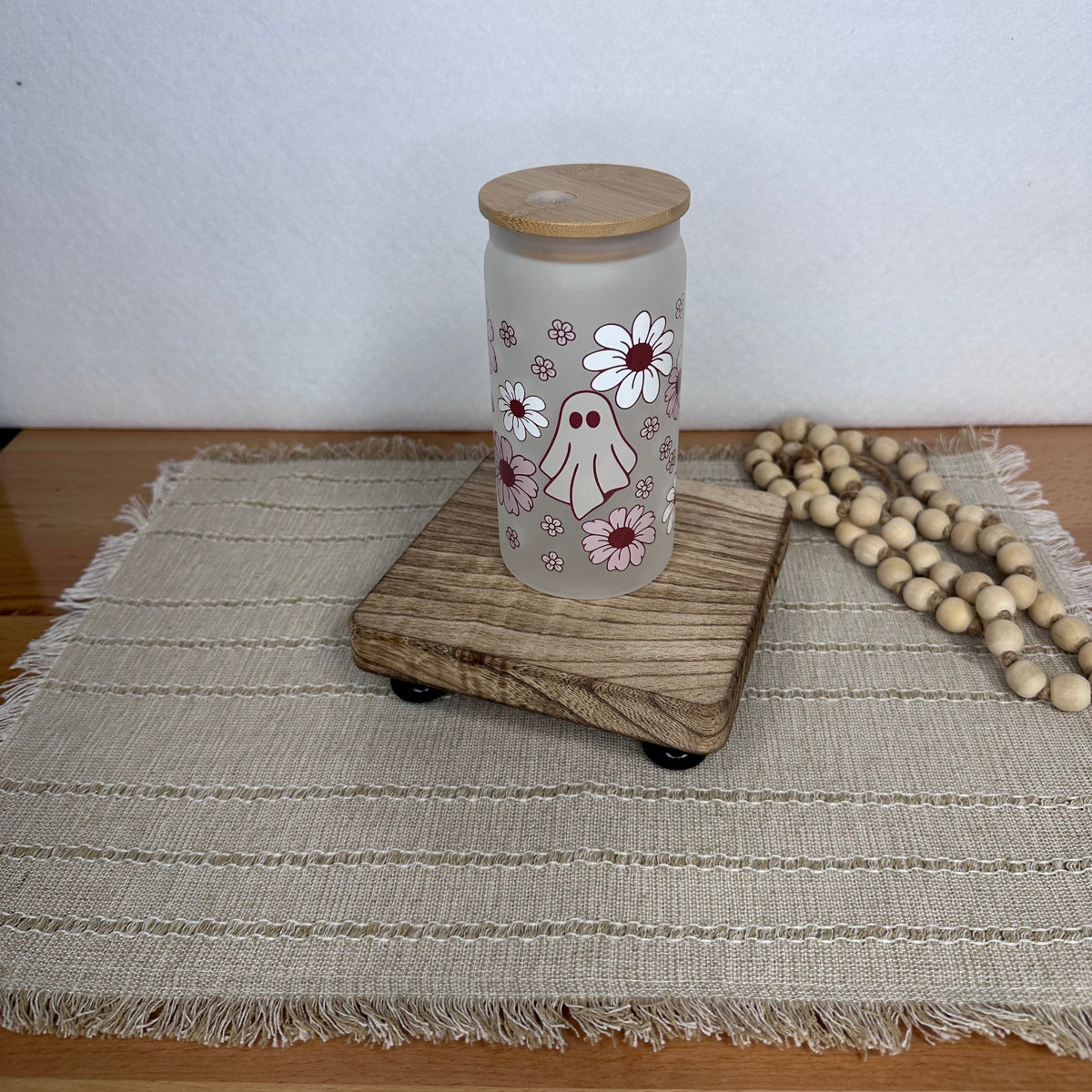A frosted glass cup with a light wood lid, decorated with a cute ghost and daisy pattern, sitting on a wooden riser on a woven placemat.