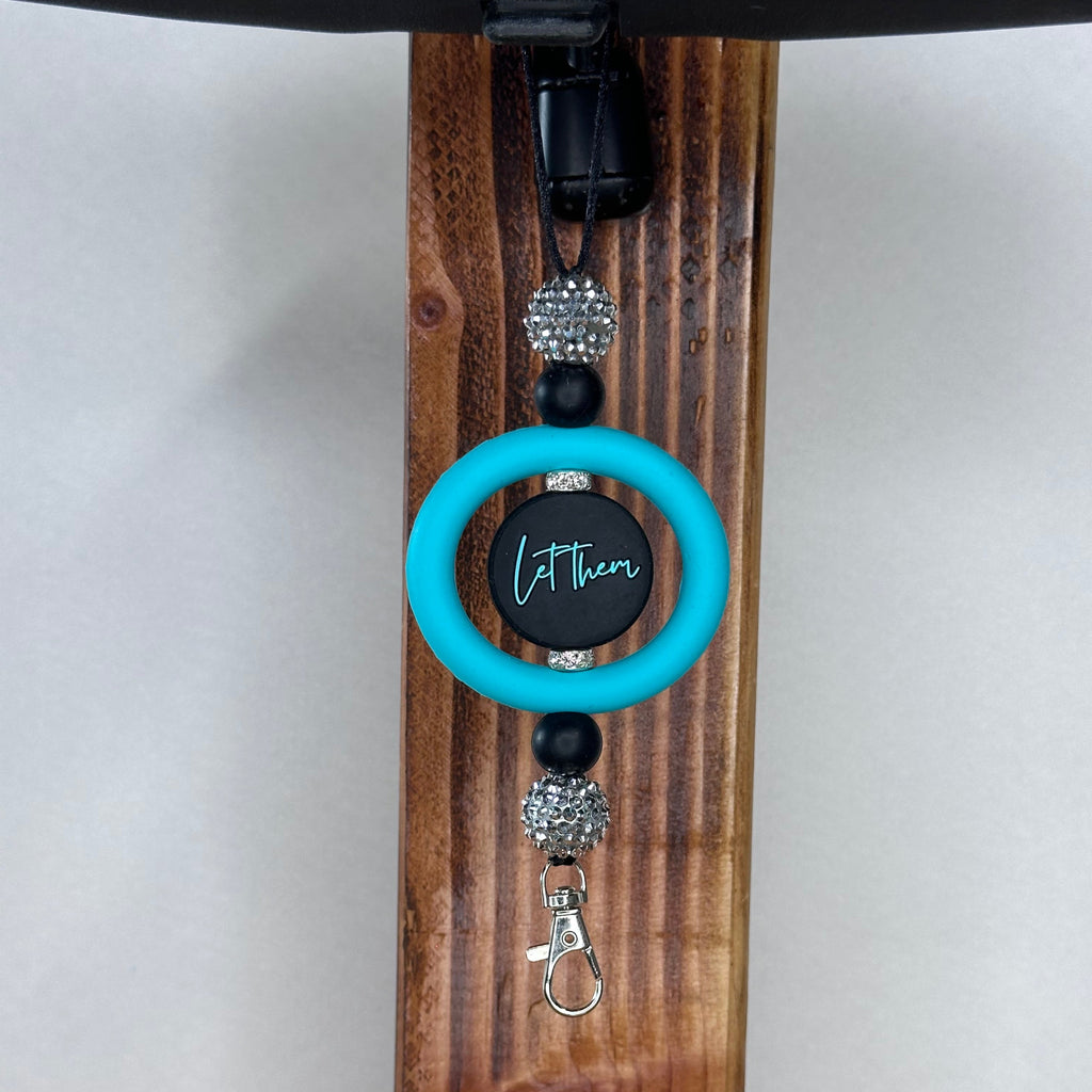 A car ornament with a bright turquoise ring, a black and turquoise  printed with 'Let them', black beads, rhinestone-embellished beads, and a silver lobster clasp, hanging on a dark wooden stand.
