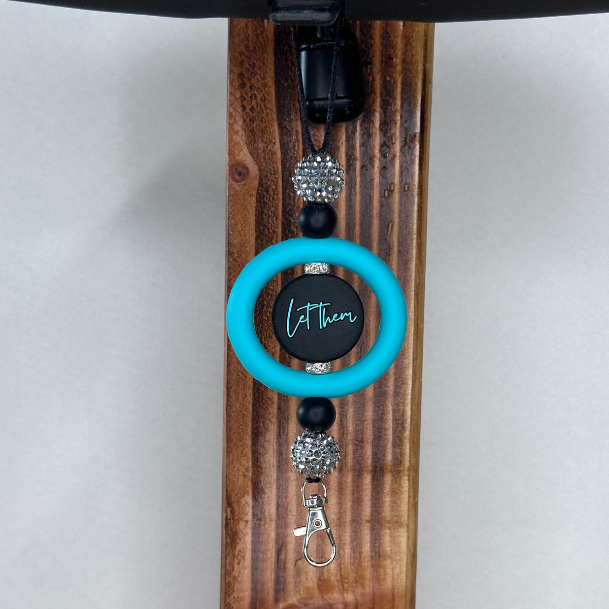 A car ornament with a bright turquoise ring, a black and turquoise  printed with 'Let them', black beads, rhinestone-embellished beads, and a silver lobster clasp, hanging on a dark wooden stand.