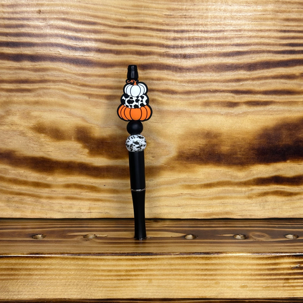 A black pen with decorative Halloween-themed bead featuring orange pumpkins, white pumpkins with black spots, and a white pumpkin, set against a rustic wood background.