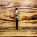 A black pen with decorative Halloween-themed bead featuring orange pumpkins, white pumpkins with black spots, and a white pumpkin, set against a rustic wood background.