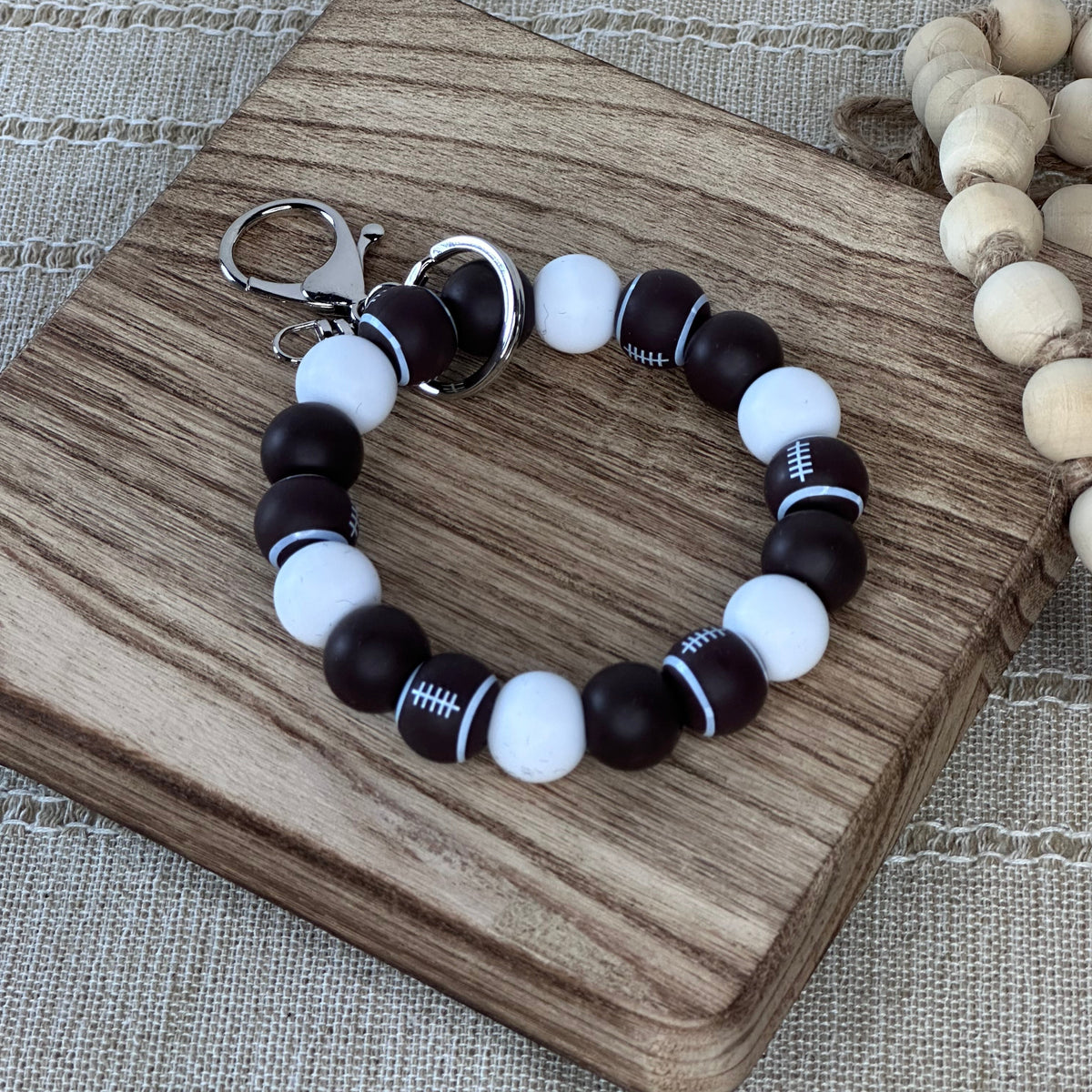 A beaded bracelet with alternating dark brown and white beads, including football-themed beads with white lacing details, displayed on a wooden block.