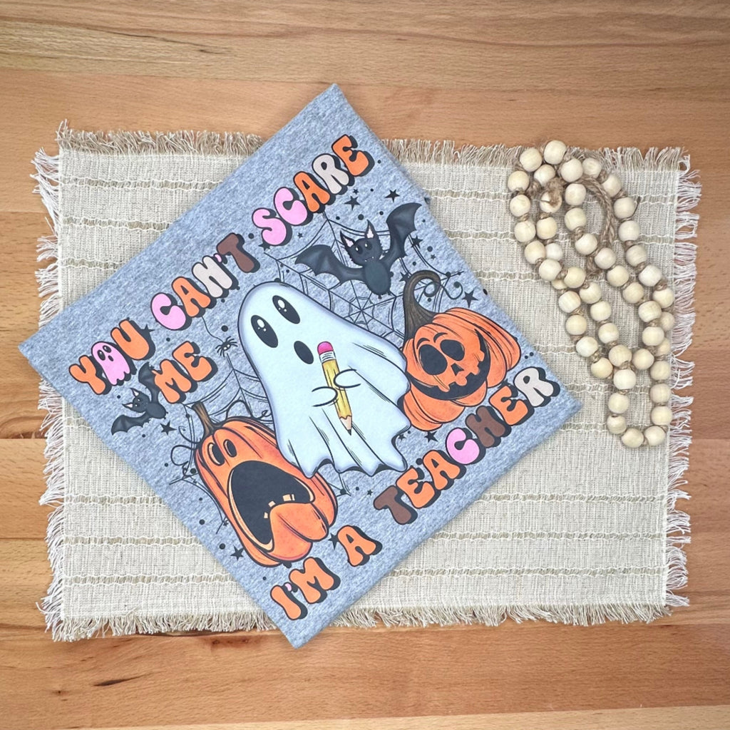 A folded Halloween-themed tshirt 'YOU CAN'T SCARE ME I'm A TEACHER' lies on a wooden table. The tshirt has a gray background with a ghost, pumpkins, bats, and text, styled with a woven placemat liner and a strand of wooden beads.