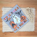 A folded Halloween-themed tshirt 'YOU CAN'T SCARE ME I'm A TEACHER' lies on a wooden table. The tshirt has a gray background with a ghost, pumpkins, bats, and text, styled with a woven placemat liner and a strand of wooden beads.