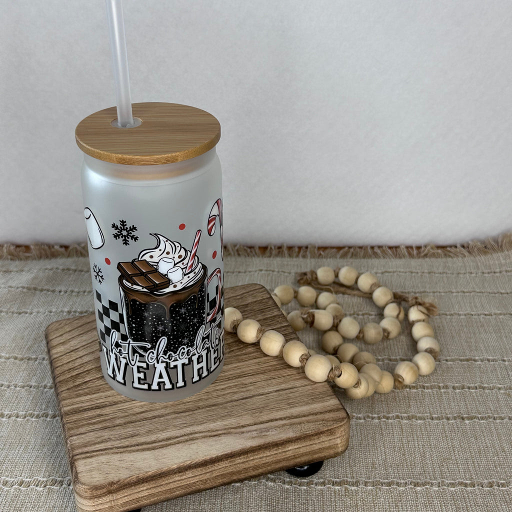 A frosted glass cup with a bamboo lid and clear straw, featuring a winter graphic that reads 'HOT CHOCOLATE WEATHER' with a hot cocoa illustration. Thecup sits on a small wooden tray next to a beaded garland against a neutral background.