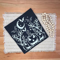 A folded black tshirt with a white Halloween print of jack-o'-lanterns, bats, and a crescent moon, displayed on a wooden table next to a wooden bead garland.