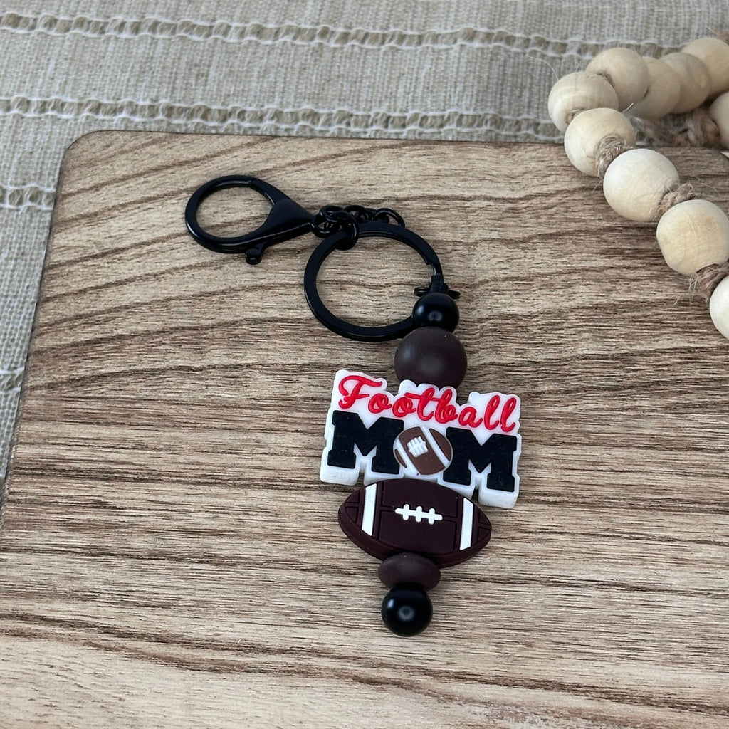 A 'Football Mom' keychain charm resting on a wooden cutting board next to a beaded necklace. The charm features a black clasp and ring, dark brown beads, a brown football, and a white and red 'Football Mom' bead.