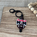 A 'Football Mom' keychain charm resting on a wooden cutting board next to a beaded necklace. The charm features a black clasp and ring, dark brown beads, a brown football, and a white and red 'Football Mom' bead.