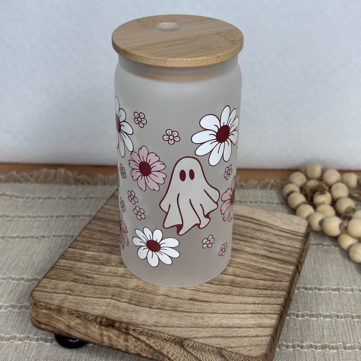 A frosted glass cup with a light wood lid, decorated with a cute ghost and daisy pattern, displayed on a wooden block.