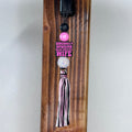 A decorative tassel charm with a pink and black striped tassel, a white rhinestone bead, a black bead, and a pink charm that reads 'SOMEBODY'S SPOILED BLUE COLLAR WIFE', hanging from a black clip on a wooden post.