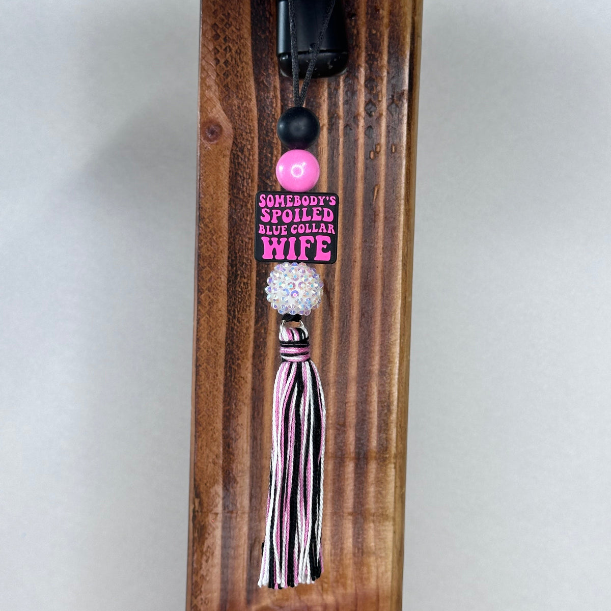 A decorative tassel charm with a pink and black striped tassel, a white rhinestone bead, a black bead, and a pink charm that reads 'SOMEBODY'S SPOILED BLUE COLLAR WIFE', hanging from a black clip on a wooden post.