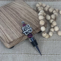 A wine stopper with a black threaded neck, a silver metal point, and a decorative charm that reads 'IT'S FINE. I'M FINE.  EVERYTHING'S FINE' with pink bead accents, displayed on a wood-grain block next to a beaded garland on a woven beige surface.