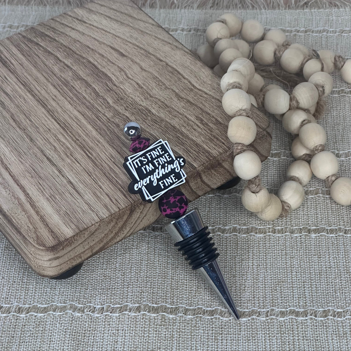 A wine stopper with a black threaded neck, a silver metal point, and a decorative charm that reads 'IT'S FINE. I'M FINE.  EVERYTHING'S FINE' with pink bead accents, displayed on a wood-grain block next to a beaded garland on a woven beige surface.