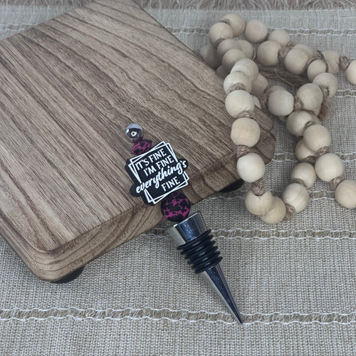 A wine stopper with a black threaded neck, a silver metal point, and a decorative charm that reads 'IT'S FINE. I'M FINE.  EVERYTHING'S FINE' with pink bead accents, displayed on a wood-grain block next to a beaded garland on a woven beige surface.
