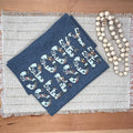 A tsirt with a white, gray, and orange patterned design reading 'SPOOKY SEASON' lies on a woven placemat on a wooden table, next to a strand of wooden beads.