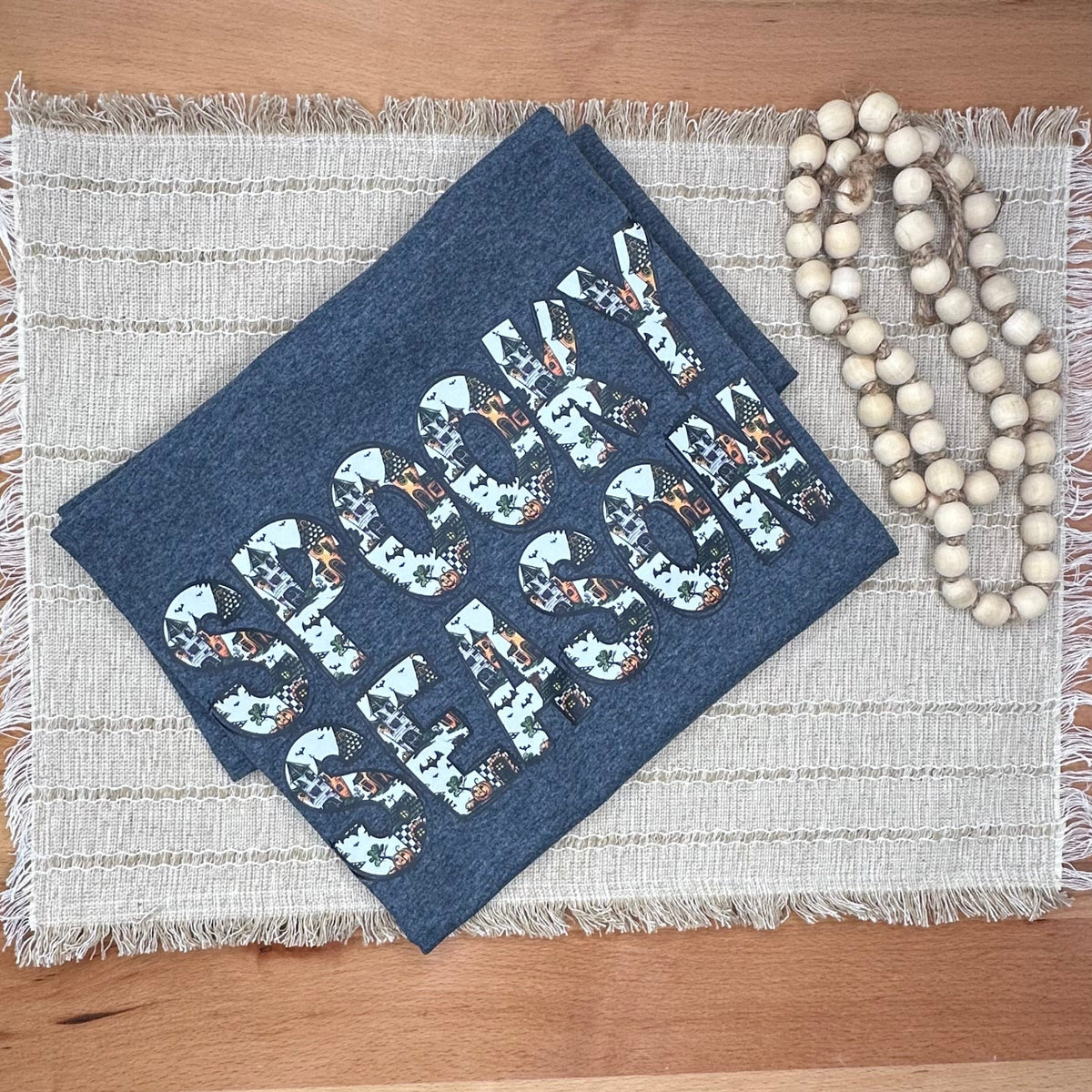A tsirt with a white, gray, and orange patterned design reading 'SPOOKY SEASON' lies on a woven placemat on a wooden table, next to a strand of wooden beads.