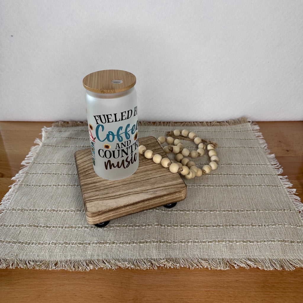 A frosted glass can cup with a bamboo lid sits on a wooden board, styled with a wooden bead garland on a woven placemat against a plain white wall.