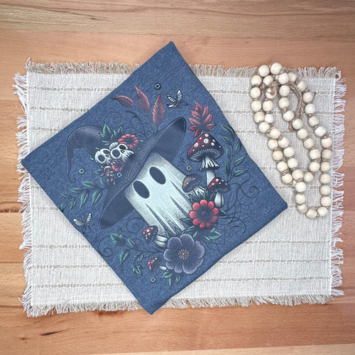 A folded tshirt with a ghost and floral design, displayed on a woven placemat on a wooden table next to a wooden bead necklace. 