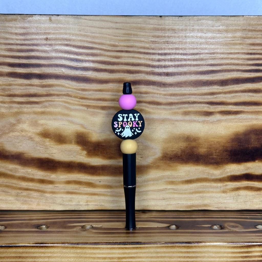 A novelty black pen with a round black and pink 'STAY SPOOKY' charm, a beige bead, and a pink bead accent, standing on a rustic wood surface.