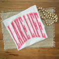 A folded white tshirt with the words 'LAKE LIFE' in bold pink collegiate-style lettering, styled on a woven placemat with a wooden bead garland on a wooden table.