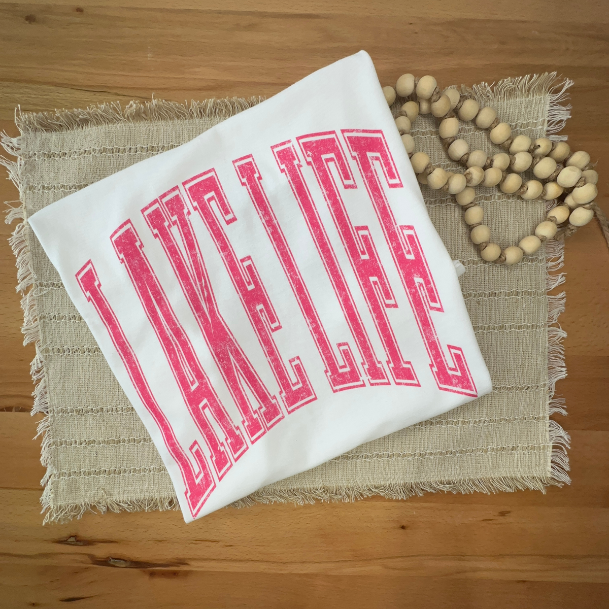 A folded white tshirt with the words 'LAKE LIFE' in bold pink collegiate-style lettering, styled on a woven placemat with a wooden bead garland on a wooden table.