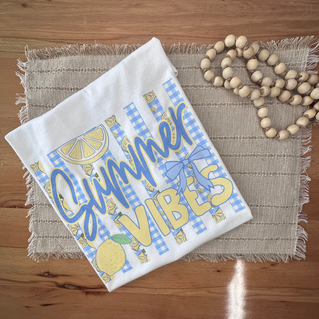 A white folded tshirt with blue and yellow 'Summer Vibes' graphic print, styled on a woven placemat with wooden beads on a wooden table.