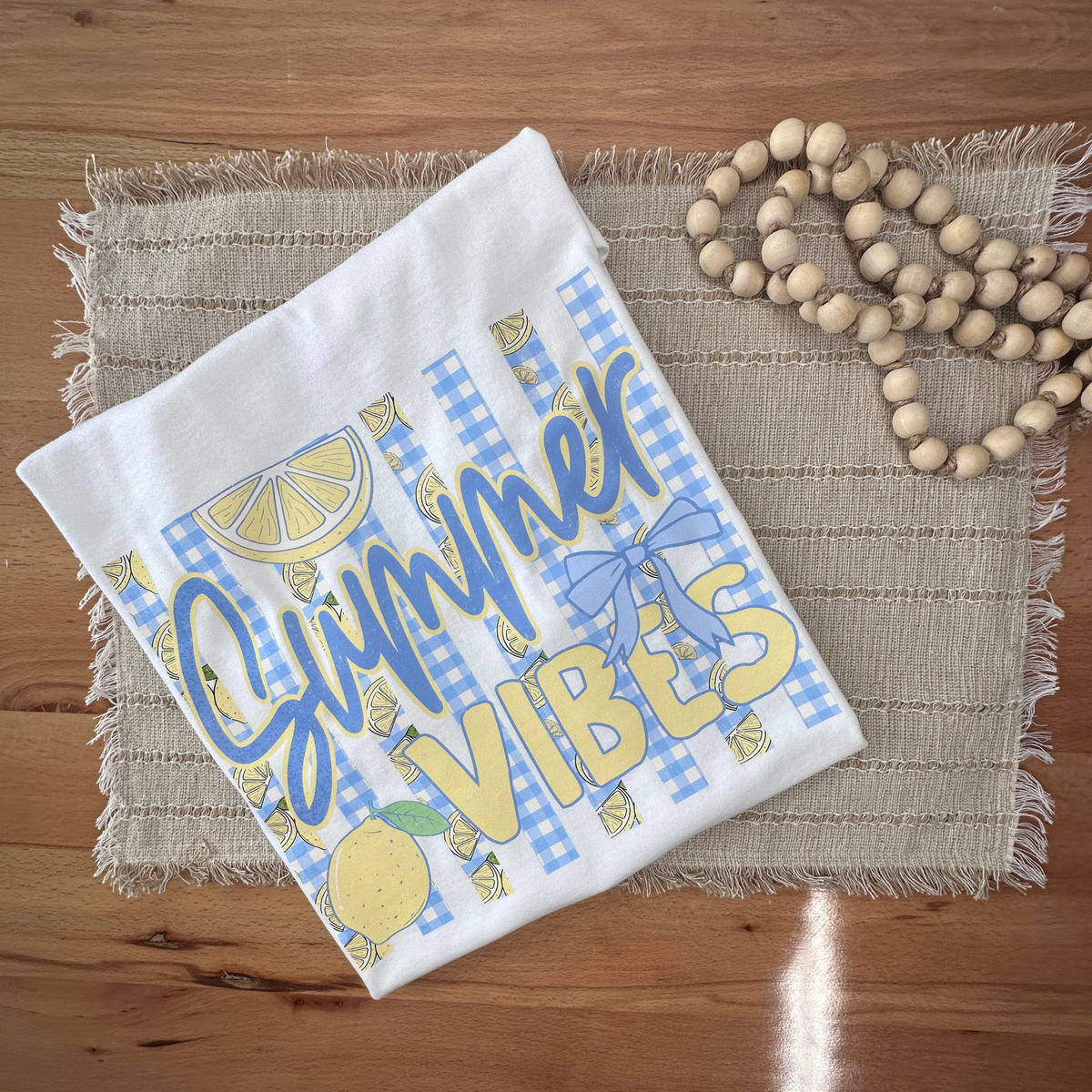 A white folded tshirt with blue and yellow 'Summer Vibes' graphic print, styled on a woven placemat with wooden beads on a wooden table.