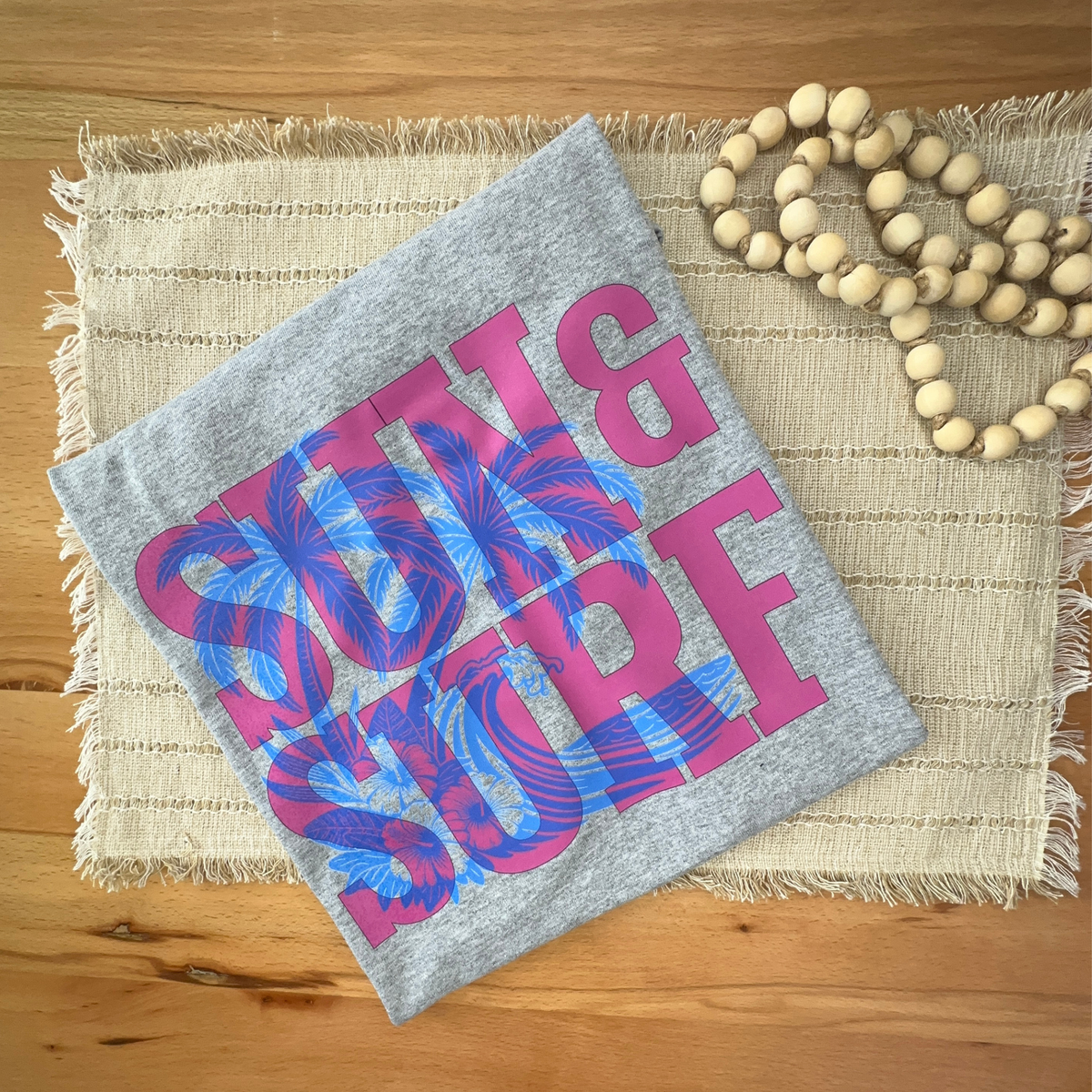 A folded heather gray tshirt featuring a bold graphic print of pink and blue palm trees and the words 'SUN & SURF'. The fabric is styled on a woven beige placemat with a wooden bead garland on a wooden tabletop.
