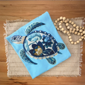 A folded light blue tshirt featuring a large faux embroidered sea turtle with blue, navy, white, and gold floral details, styled on a woven placemat with wooden beads on a wood tabletop.