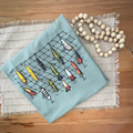 A folded tshirt in pale green, with a fishing net and colorful fishing lures design, styled flat-lay on a woven placemat with wooden beads on a wooden table.
