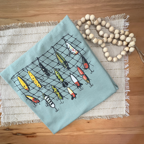 A folded tshirt in pale green, with a fishing net and colorful fishing lures design, styled flat-lay on a woven placemat with wooden beads on a wooden table.